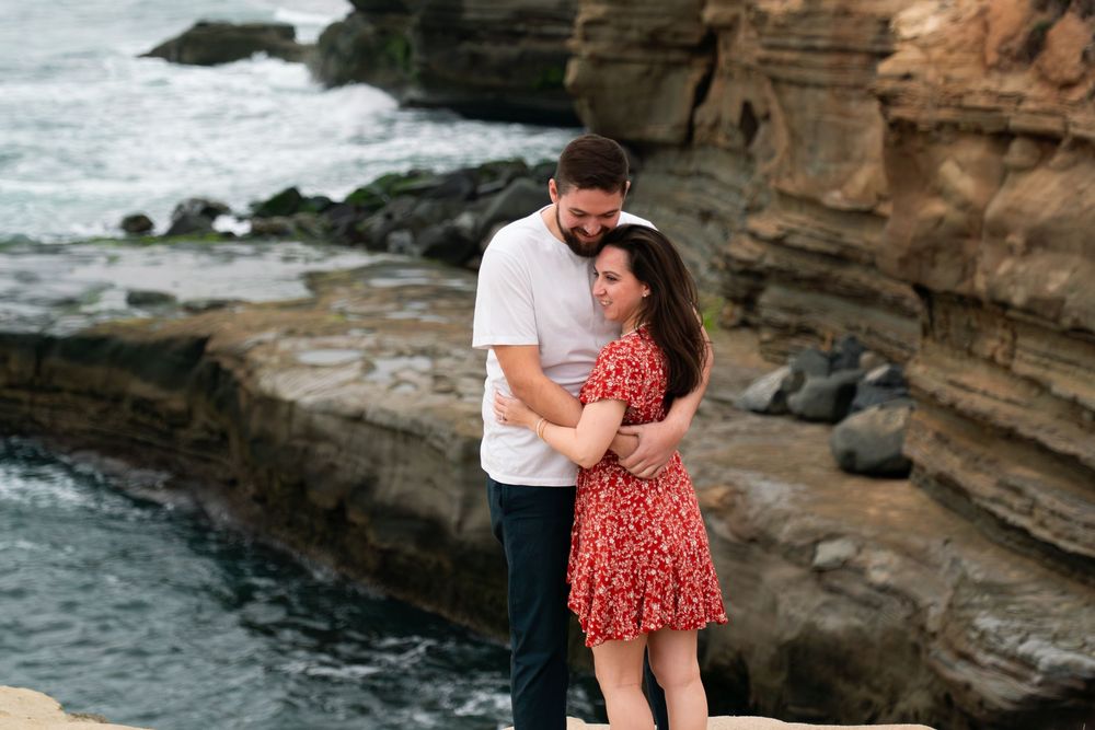 Two people embrace romantically on rocky coastal cliffs overlooking the ocean at sunset.