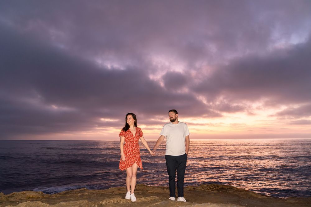 A couple stands hand in hand on a seaside cliff during a dramatic purple and orange sunset.