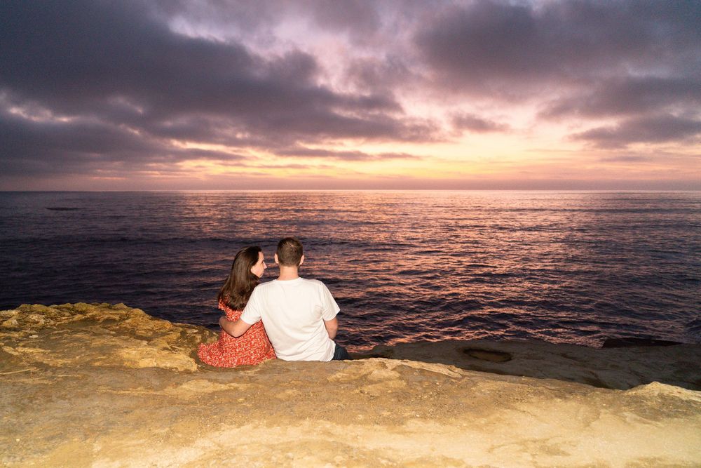 Two people sit together on a coastal cliff watching a dramatic sunset over the ocean horizon.