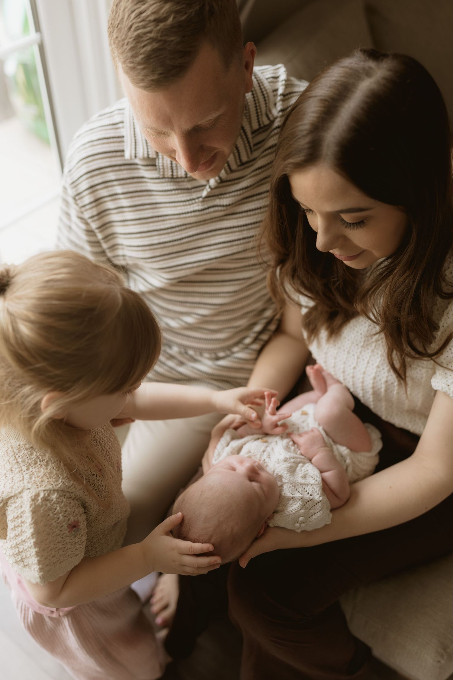 Family gathered on couch sharing tender moment with newborn baby while toddler sibling looks on in soft natural light.