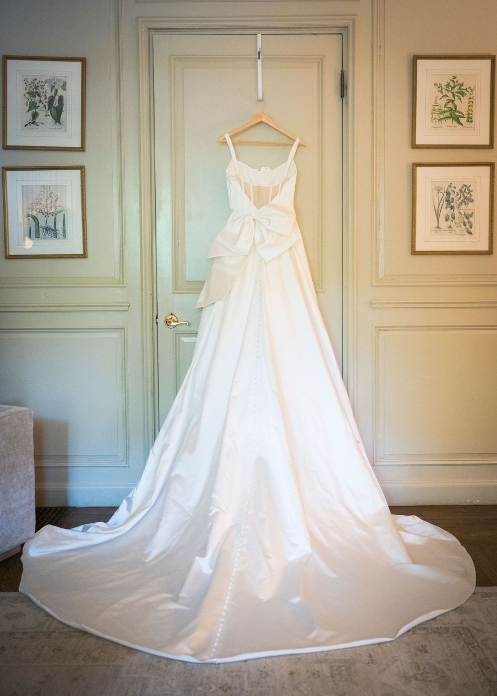 An elegant white wedding gown hanging on display against a pale wall with framed botanical prints.