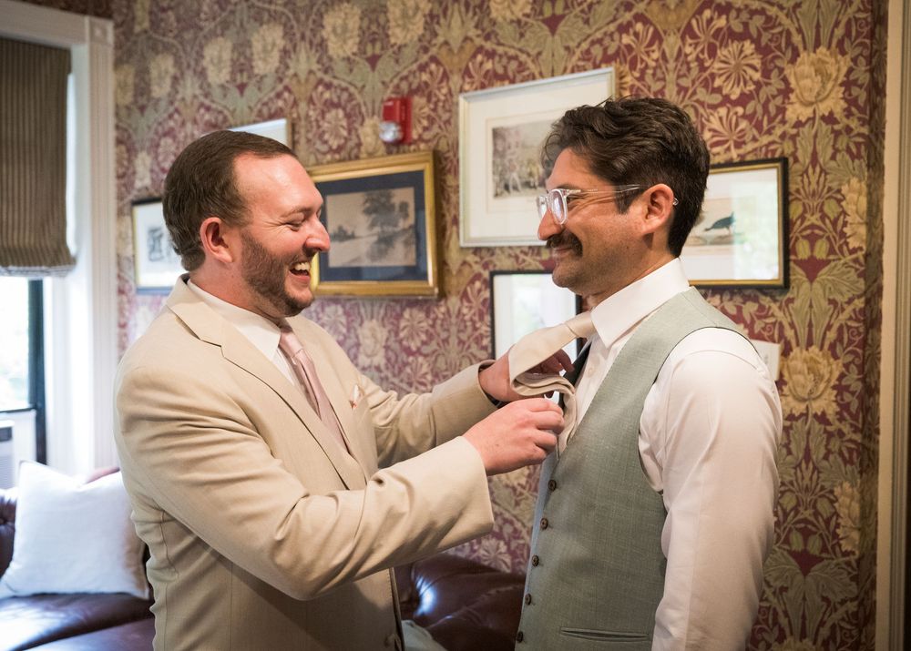 Two well-dressed men share a lighthearted moment in front of a wallpapered wall with antique frames.