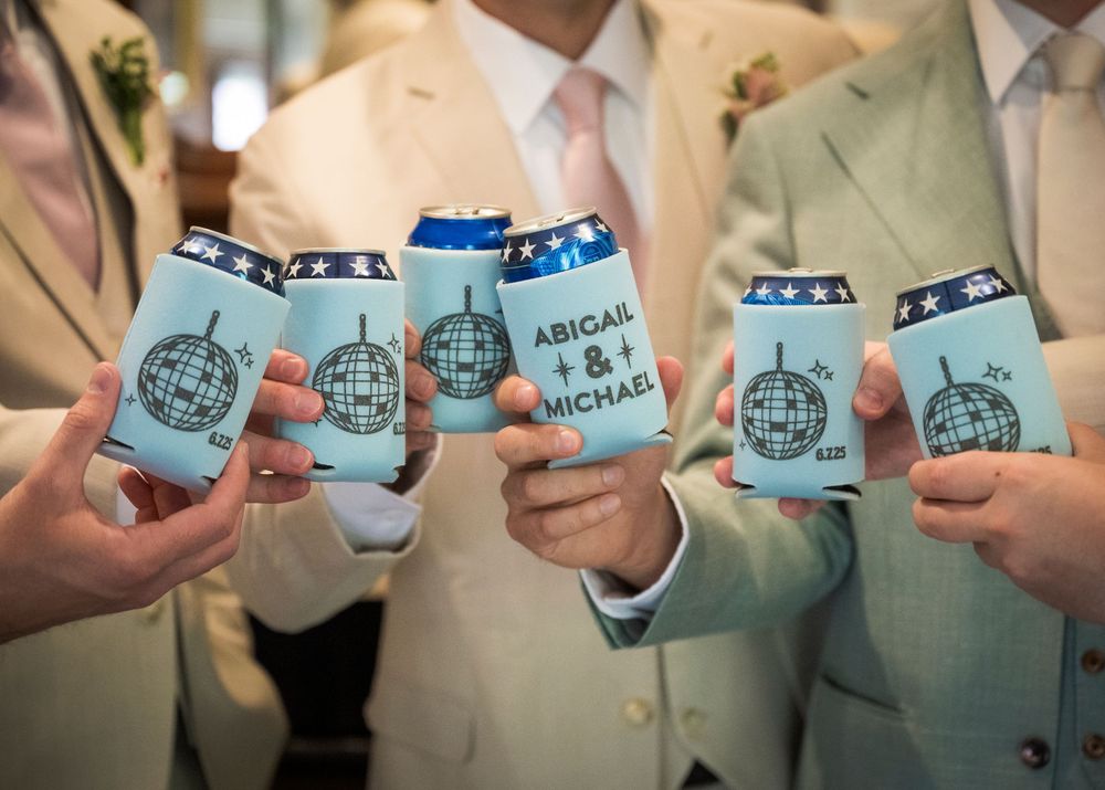 Groomsmen holding matching light blue koozies with custom logo designs at a wedding celebration.