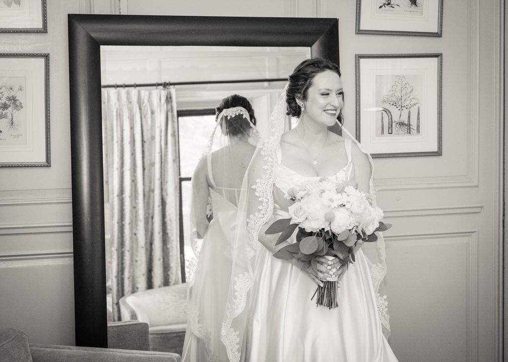 A black and white photograph captures someone admiring their wedding dress reflection while holding a flower bouquet.