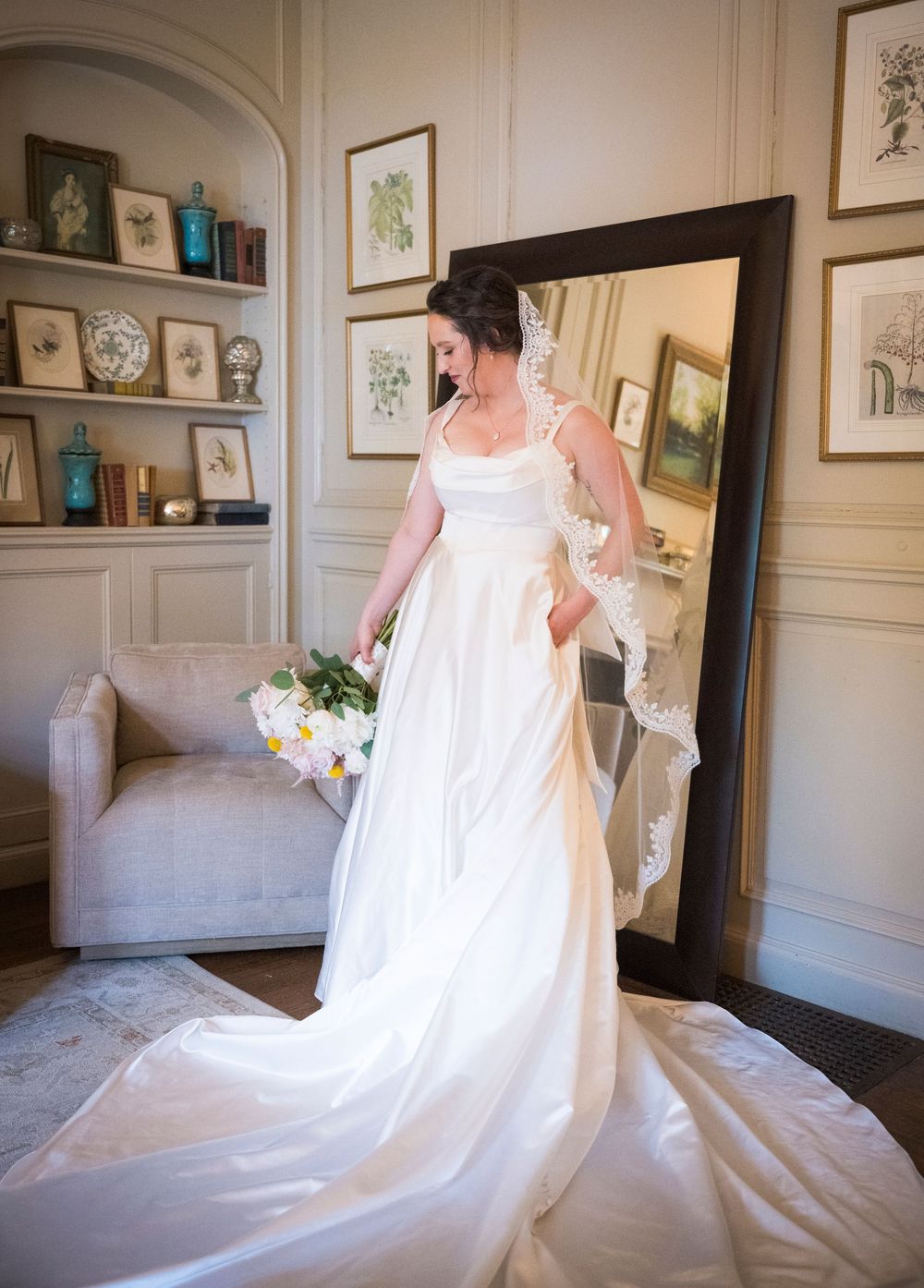 Bride in a flowing white wedding gown looks at her reflection in an elegant room with framed art and built-in shelving.