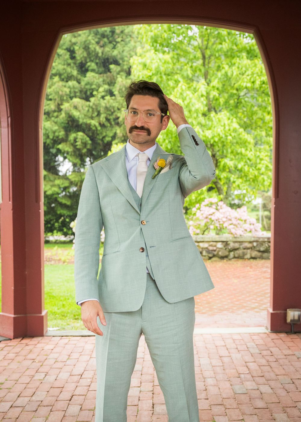 A groom in a mint green suit poses under a brick archway during a wedding photo session.