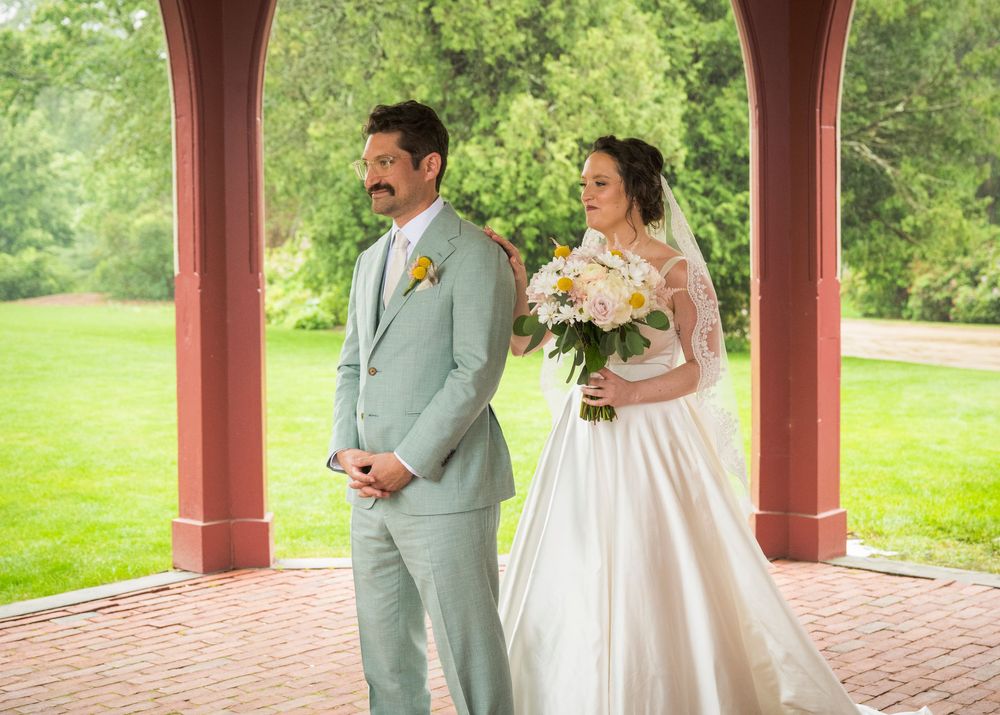 A couple shares their first look moment under a brick gazebo in a lush garden setting.