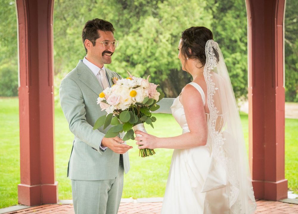 A bride and groom share their first look during an outdoor wedding ceremony under a red gazebo with lush greenery.