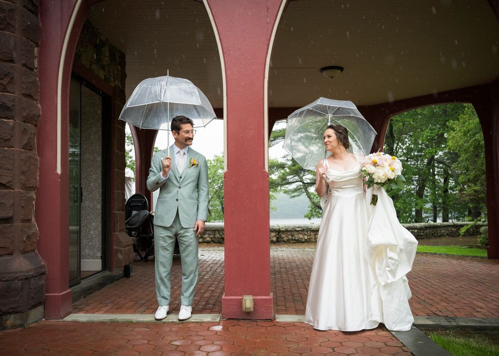 A couple shelters under umbrellas while posing beneath red archways on their rainy wedding day.