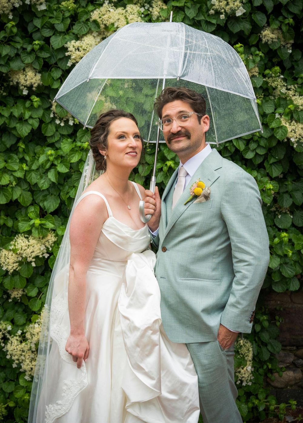 A couple shares an intimate moment under a clear umbrella against a backdrop of flowering bushes.