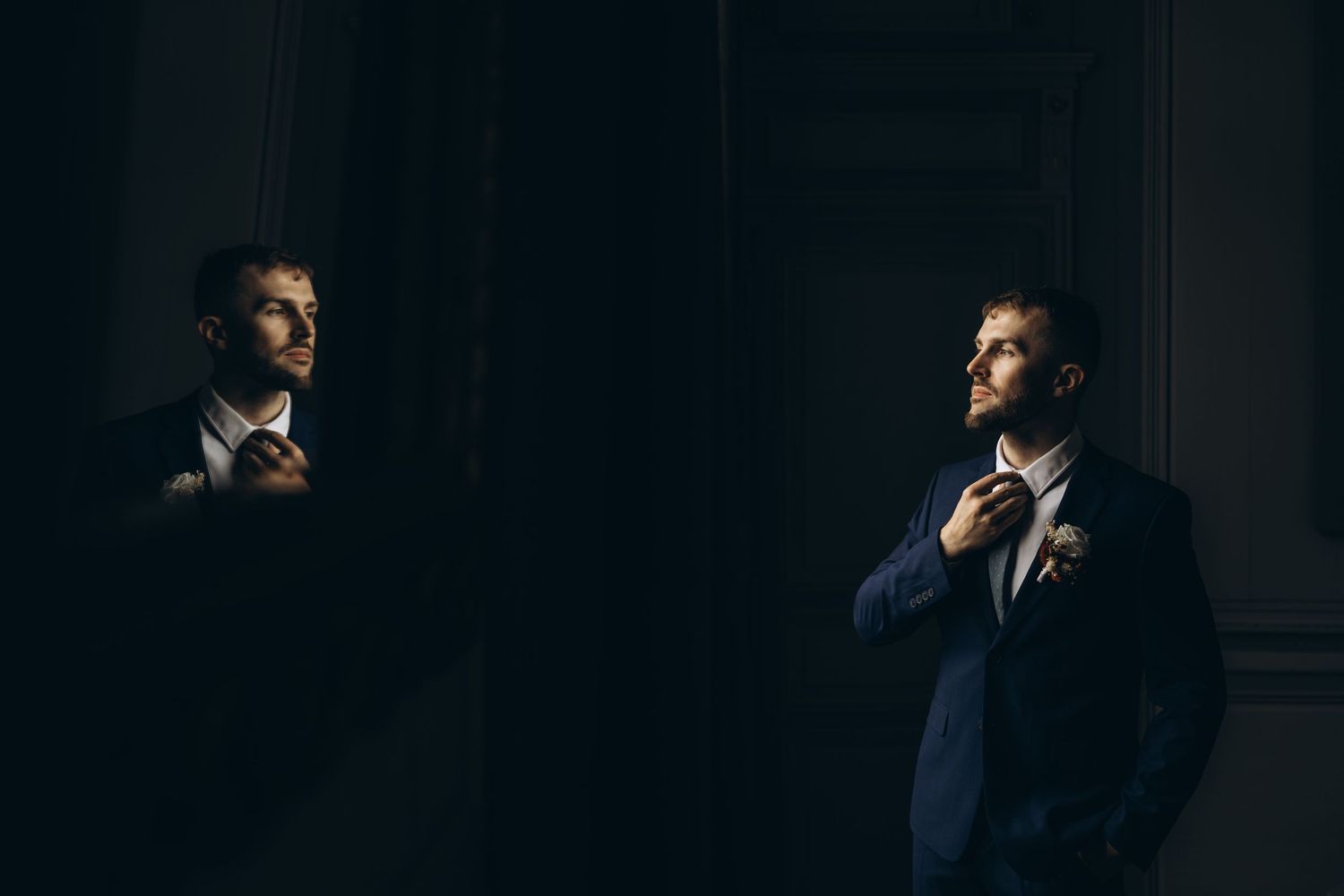 Groom in dark suit adjusts tie in moody mirror reflection with dramatic lighting.