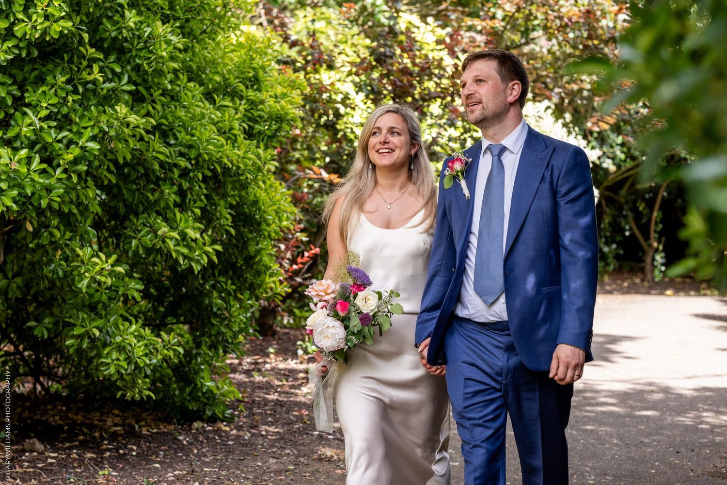 Happy couple strolls along garden path with bride holding pink bouquet and groom wearing navy suit.