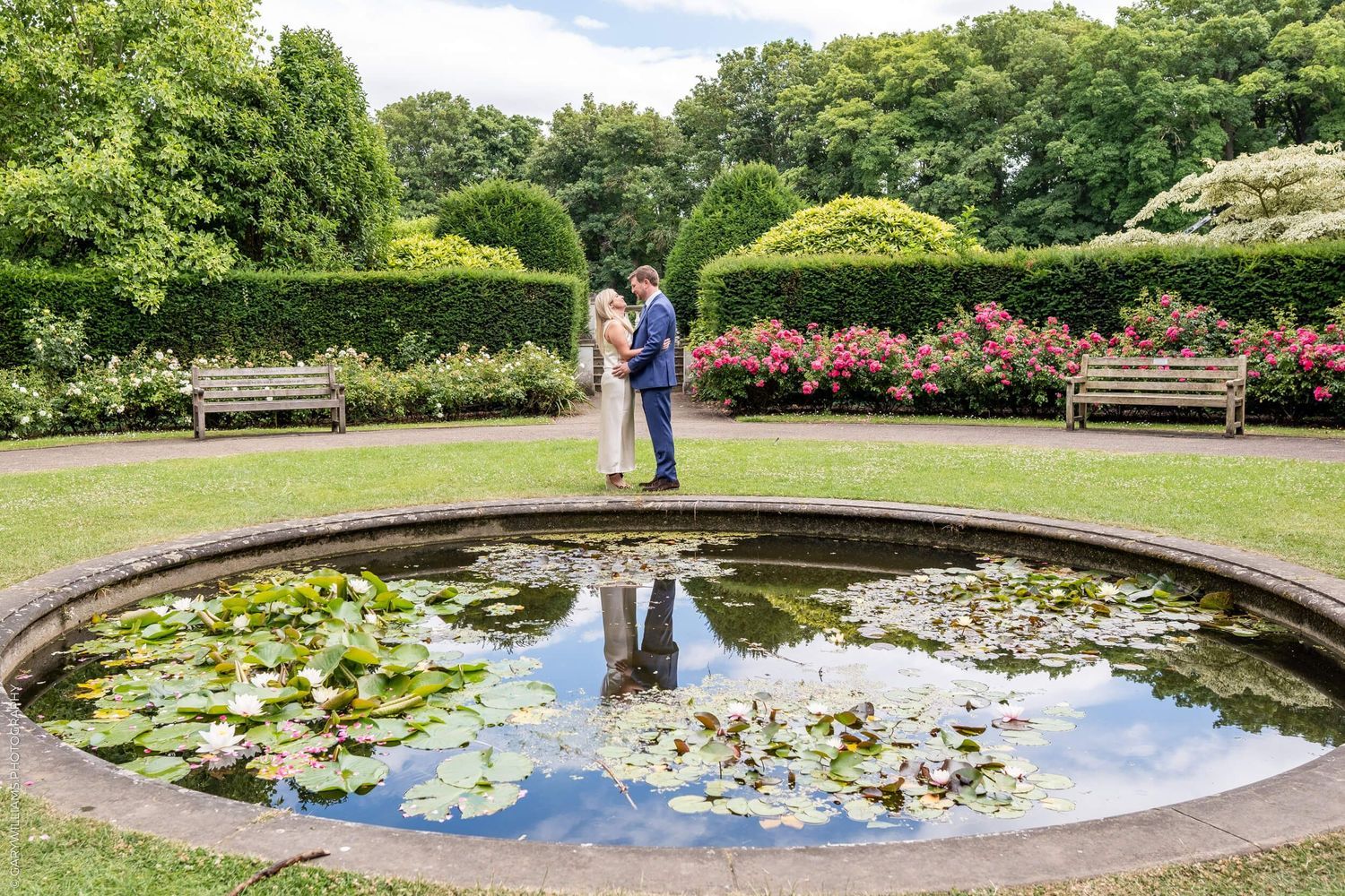 A couple stands together beside a circular lily pond with benches and landscaped gardens in the background.