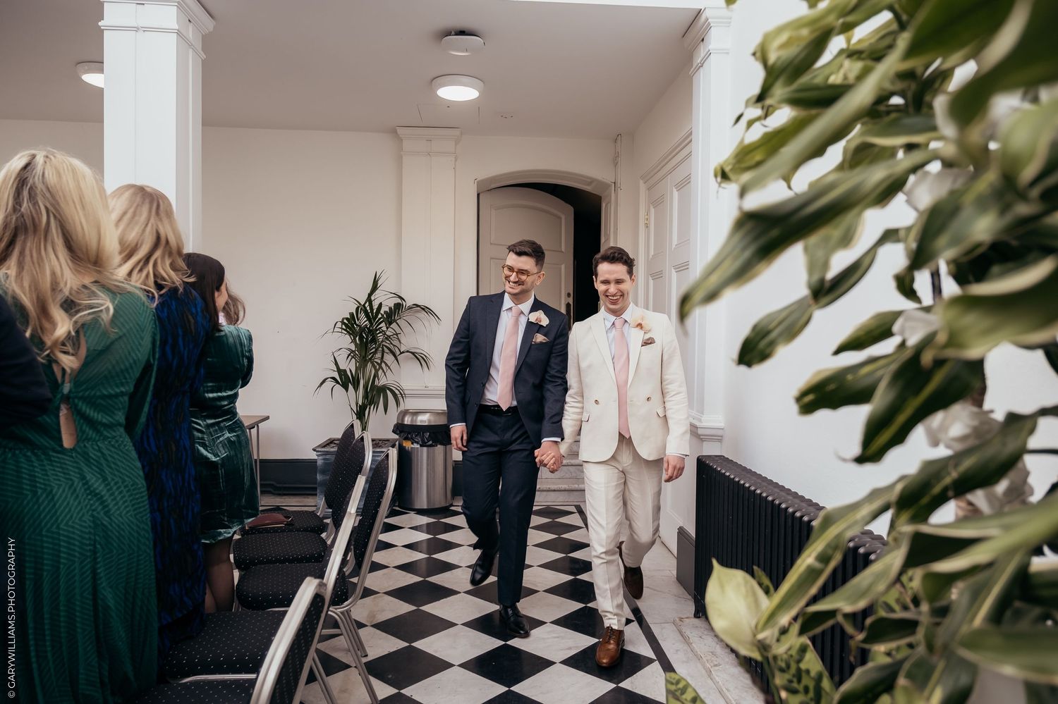 Wedding couple walks down a black and white checkered hallway as guests look on, with lush greenery visible.