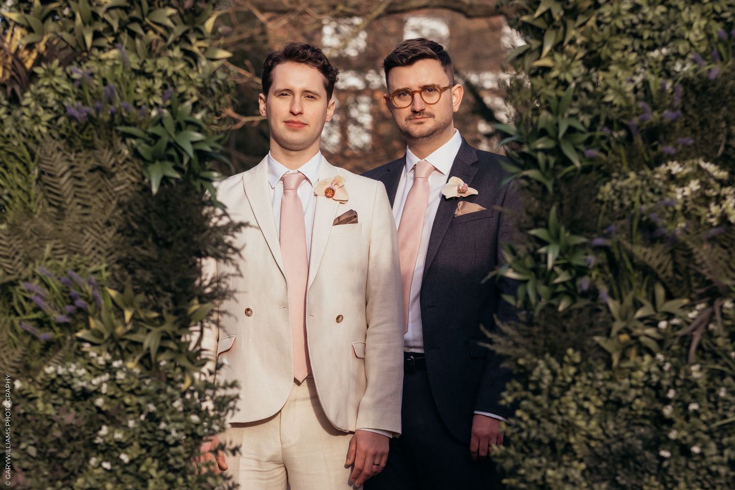 Two people in coordinated wedding attire stand together outdoors with greenery in the background.