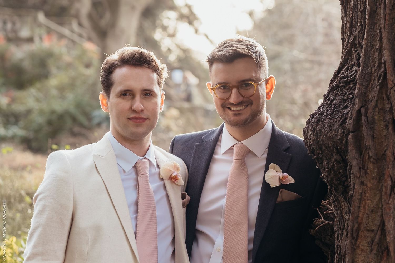 Two formal-dressed people in suits and peach-colored ties smile together near a tree at an outdoor event.