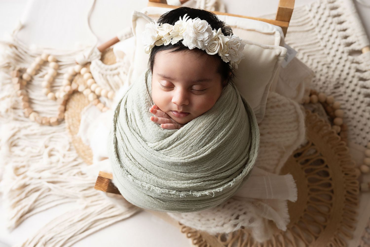 Sleeping newborn wrapped in sage green swaddle wearing floral crown on textured backdrop.