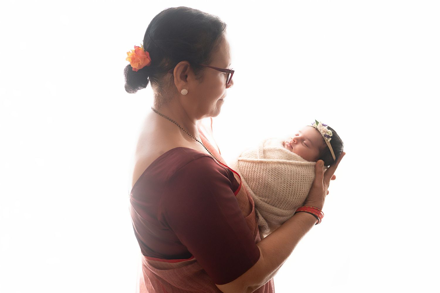 Woman in burgundy dress holding newborn baby wrapped in cream blanket against bright white background.