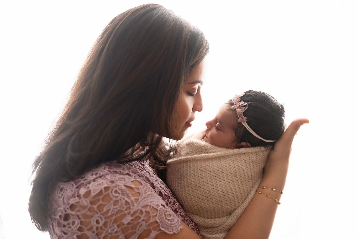 Mother in pink dress snuggling newborn baby wrapped in beige blanket against white background.