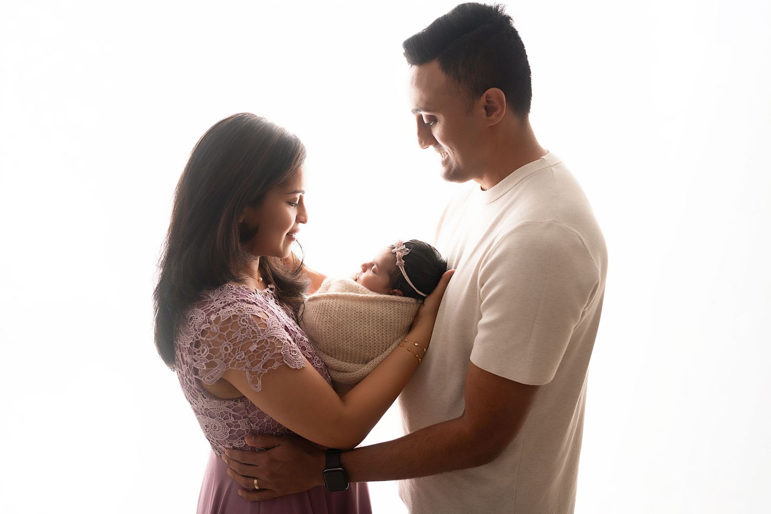 Parents sharing tender moment with newborn baby wrapped in beige blanket against white backdrop.