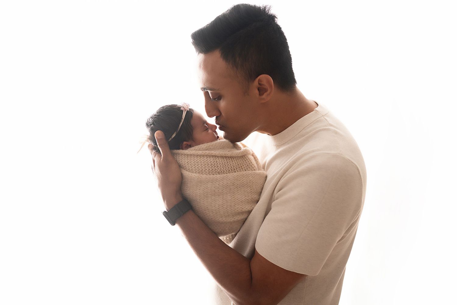 Parent gently kisses newborn baby wrapped in beige blanket against white backdrop.