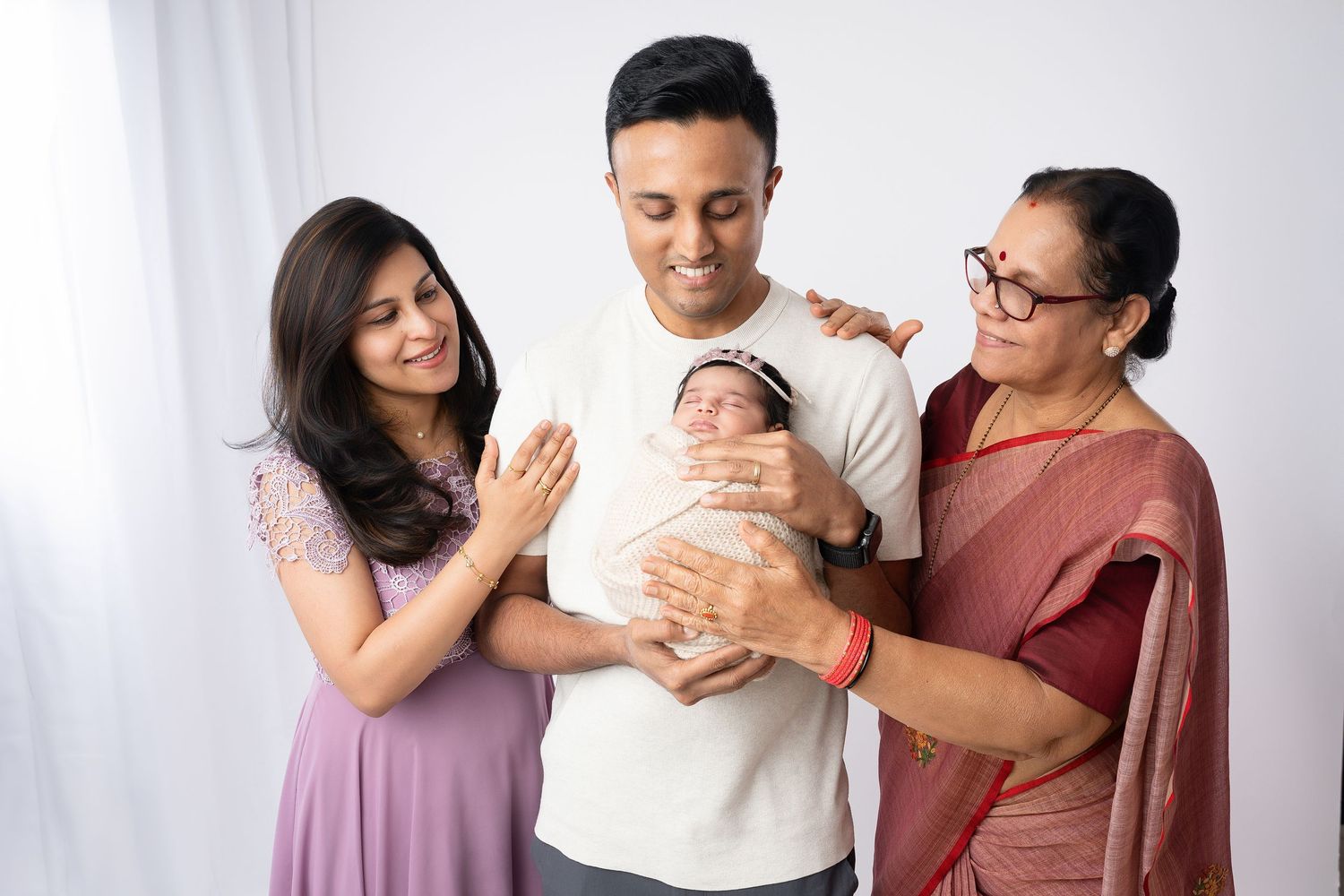 Three generations gather around newborn baby in white studio setting.