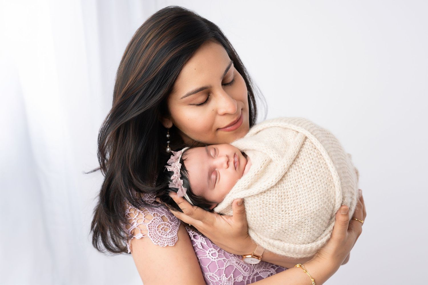 Mother in lavender dress embraces sleeping newborn in cream wrap against white background.