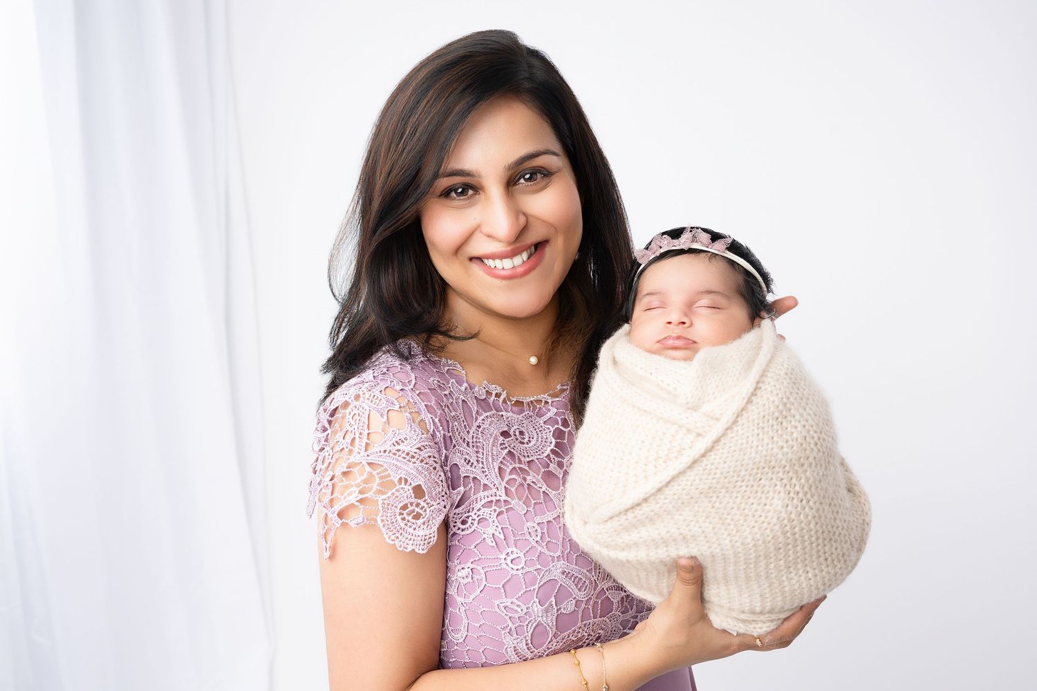 Woman in purple lace dress holds sleeping newborn wrapped in cream blanket.