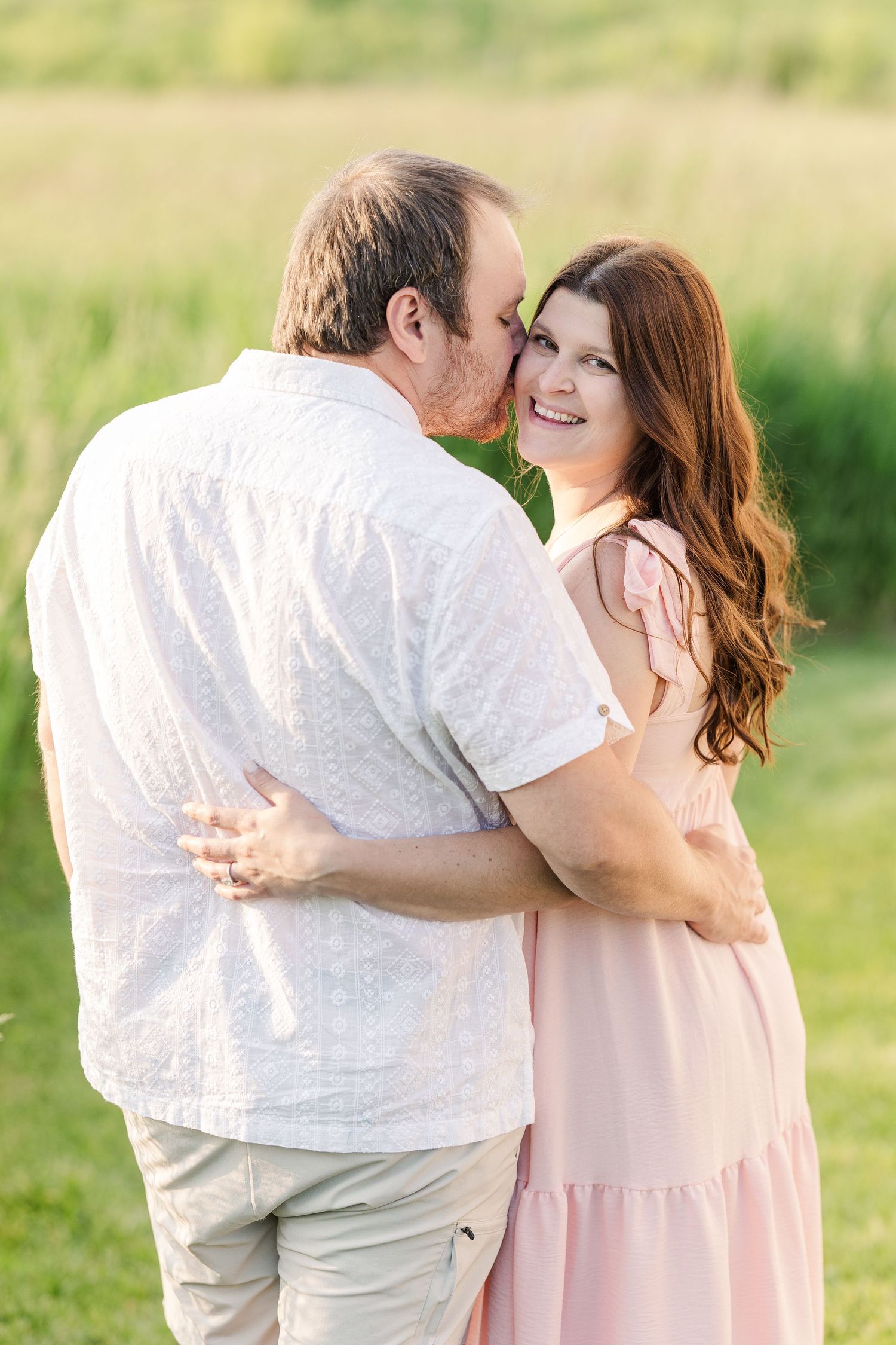 Two people embracing outdoors in a grassy field with soft lighting and wearing light colored clothing.