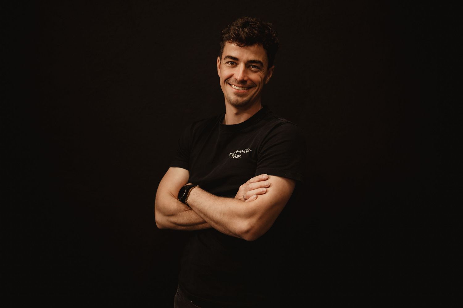 A person in a black t-shirt stands with crossed arms against a dark background, smiling warmly at the camera.