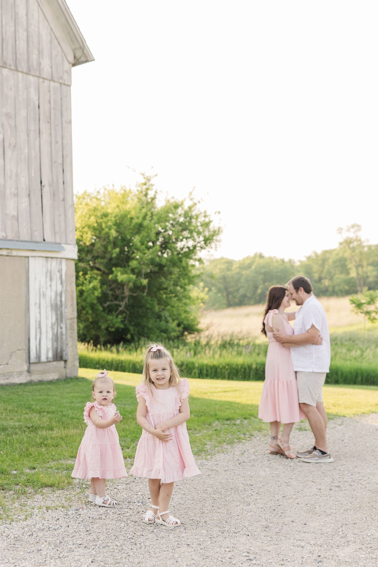 Two young girls in matching pink dresses play on path near rustic barn while couple embraces in background.