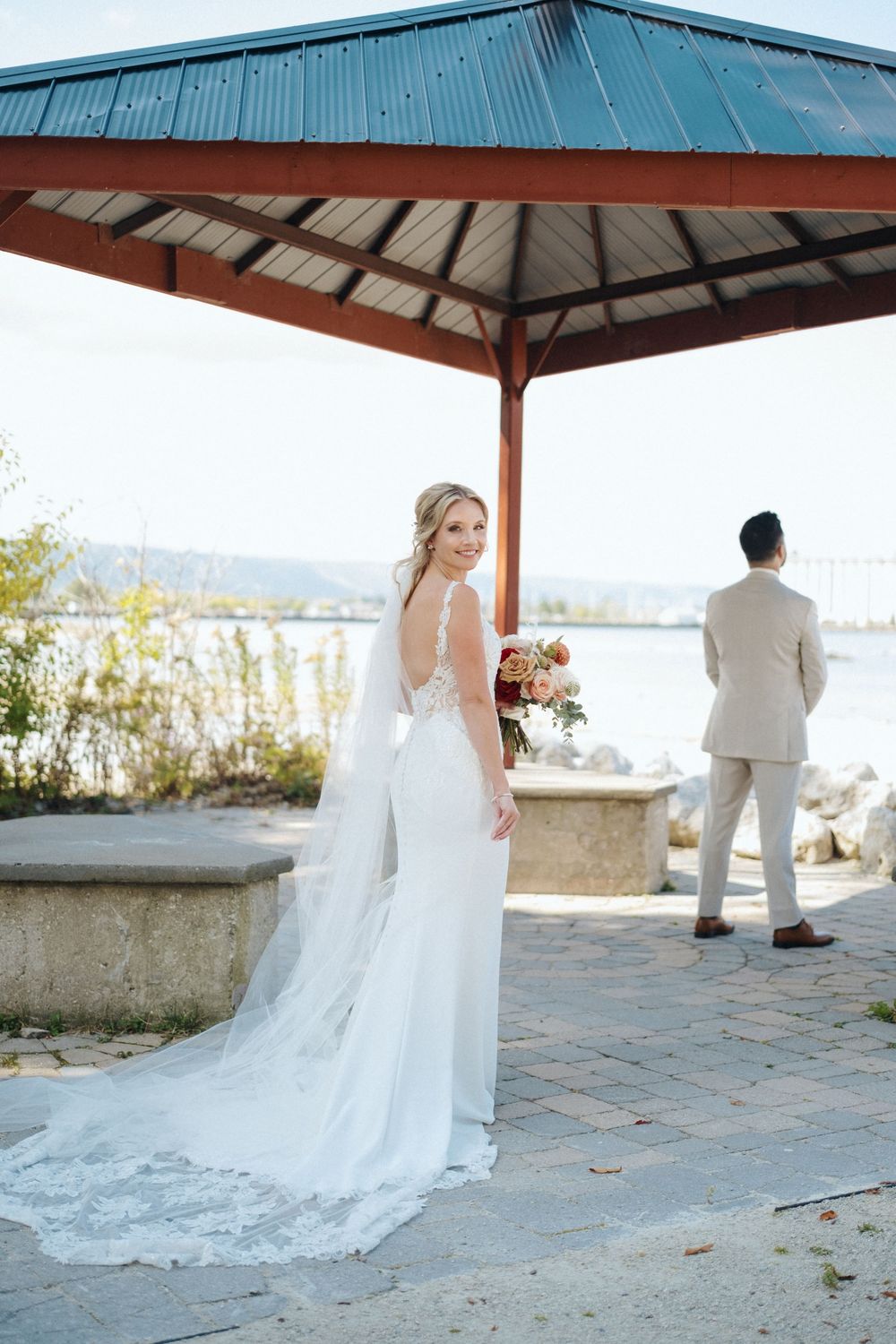 Bride in elegant white dress poses while groom waits under waterfront pavilion.