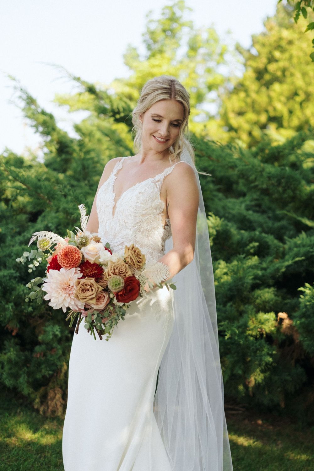 Bride holds a lush bouquet with coral and cream flowers against a green garden background.