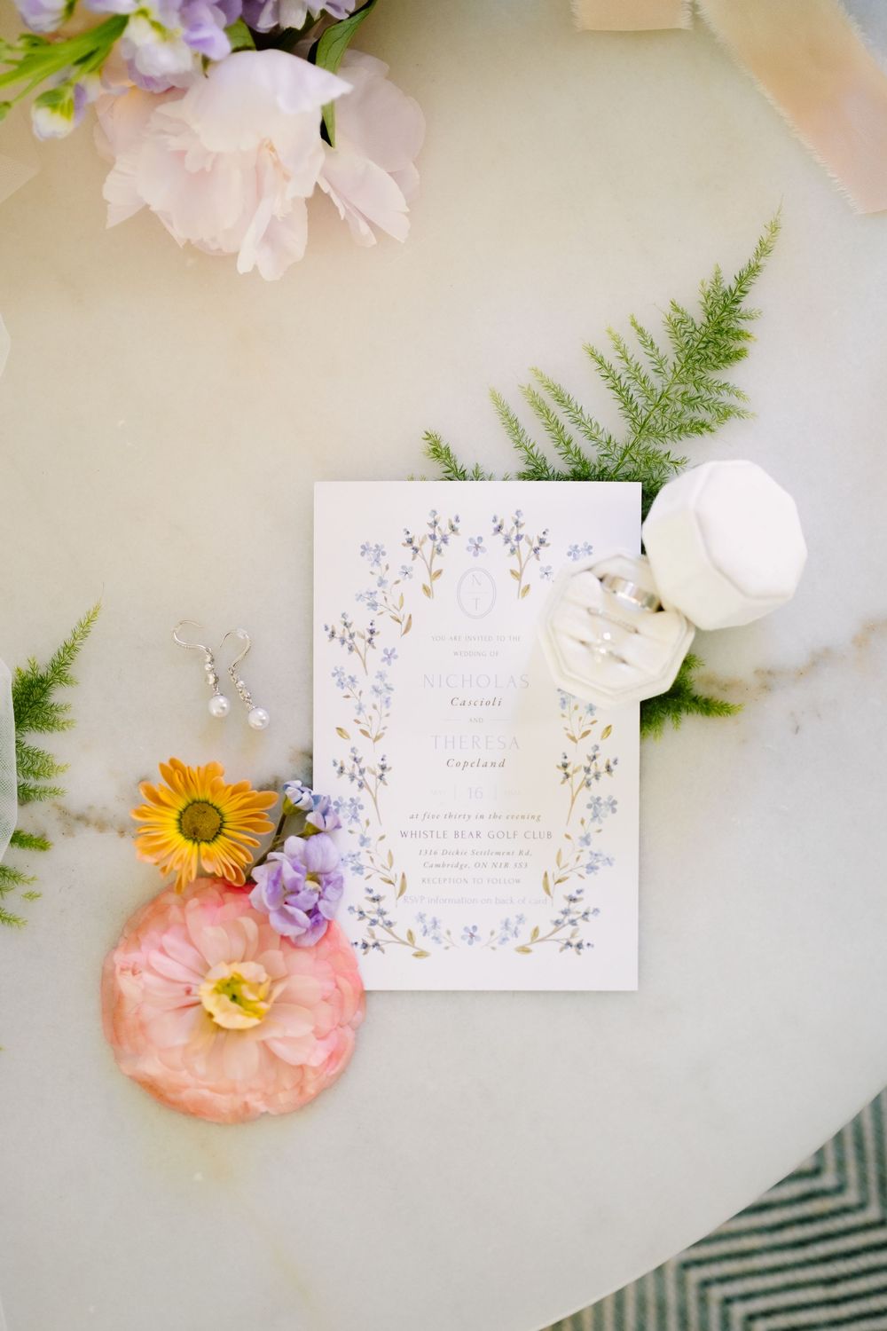 Wedding invitation with delicate floral design surrounded by fresh flowers and ferns on light background.