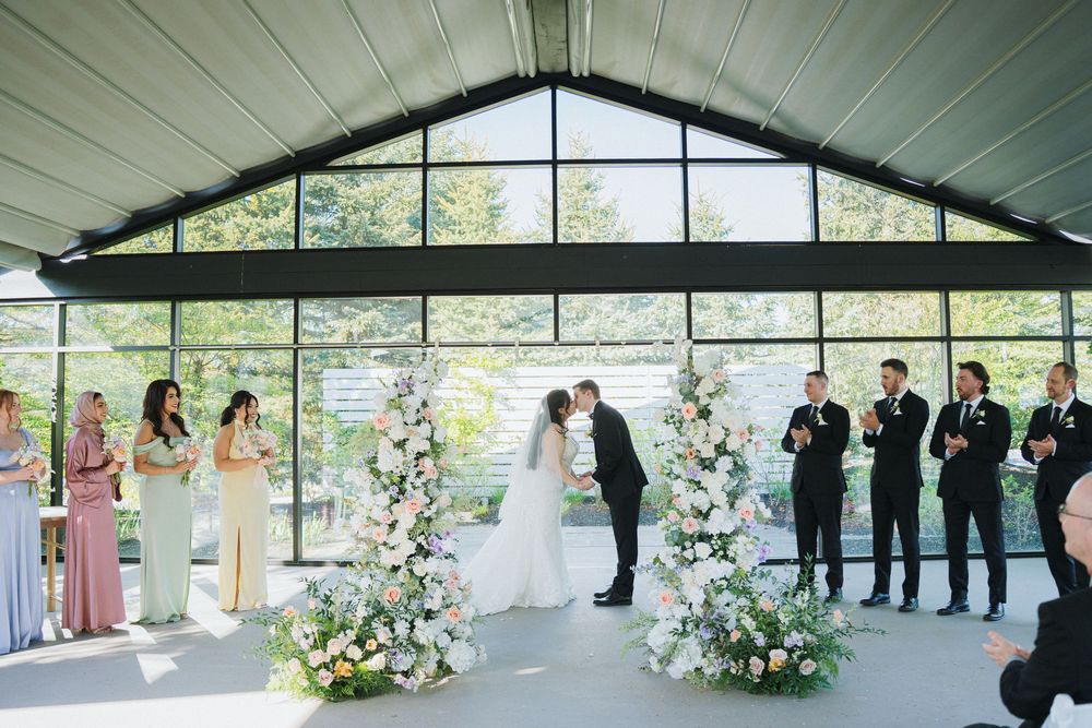 A wedding ceremony takes place in a modern glass-enclosed venue with floral arrangements and bridesmaids in pink dresses.