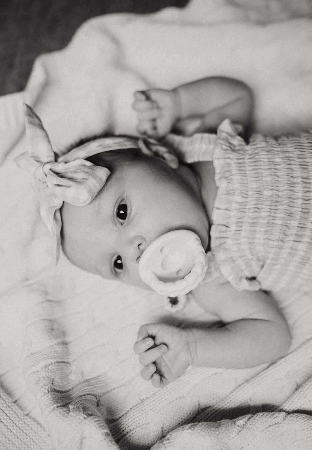 Sweet black and white photo of a baby with a pacifier lying on soft textured blankets.
