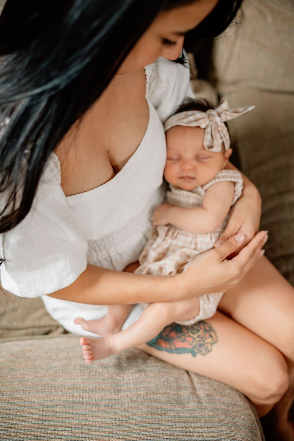 Parent in white outfit holding sleeping baby wearing bow headband and floral outfit on couch with visible tattoo artwork.
