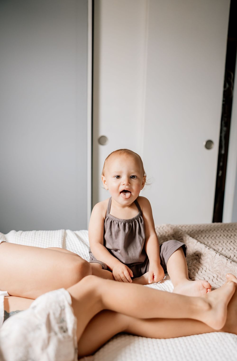 Small baby in brown top and gray shorts sits on white bedding at home.