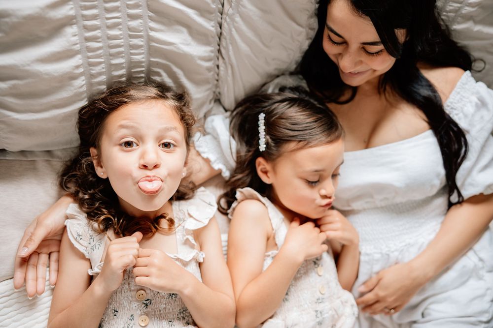 Three people laying on white bedding, with one playfully sticking out their tongue while another appears to be sleeping.