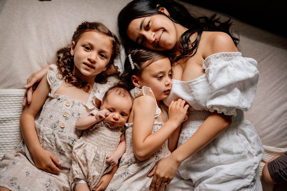 A tender moment captured of a family cuddling together on white bedding, wearing light colored dresses.