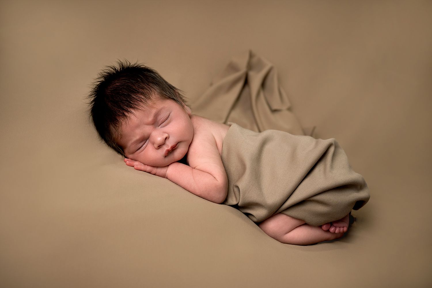 A peaceful sleeping newborn baby wrapped in a tan blanket on neutral beige background in studio lighting.