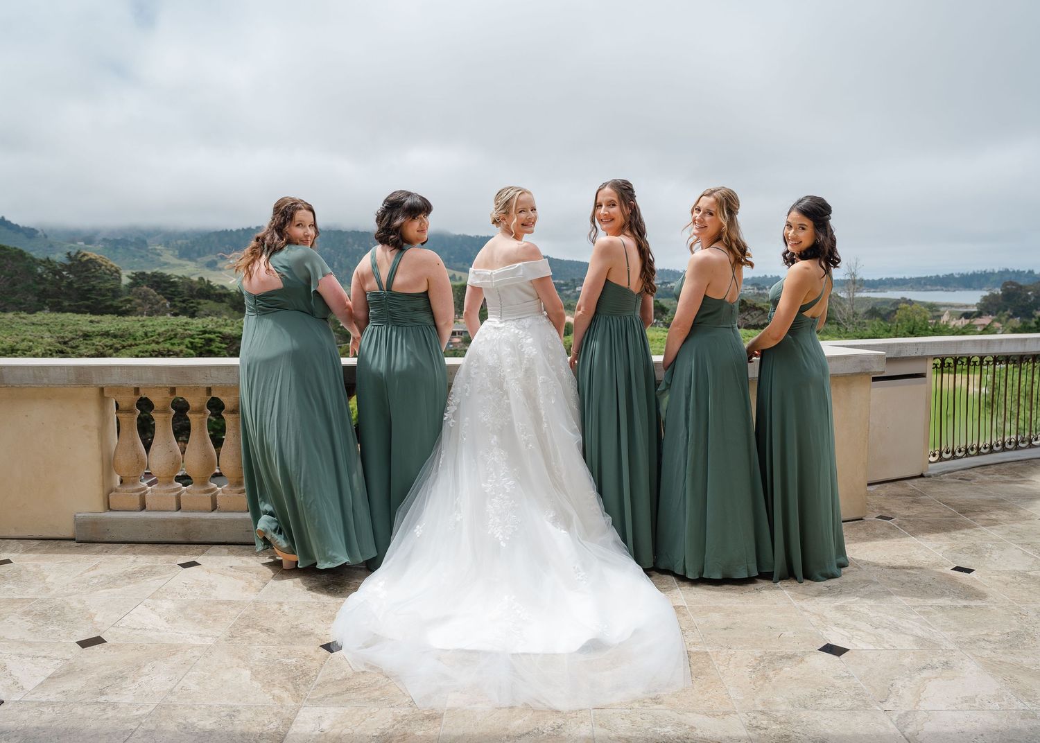 A wedding party in sage green bridesmaid dresses laughs together on an outdoor balcony overlooking rolling hills.