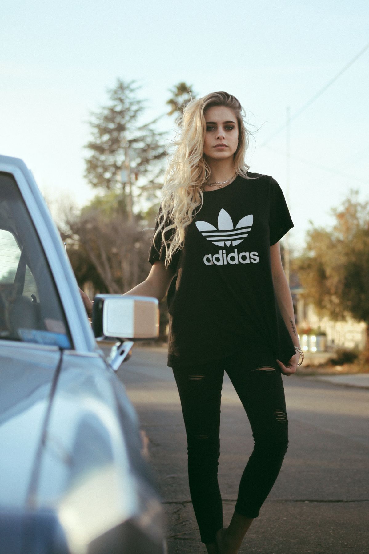 Athletic wear fashion shot featuring a black Adidas t-shirt and skinny jeans next to a parked car.