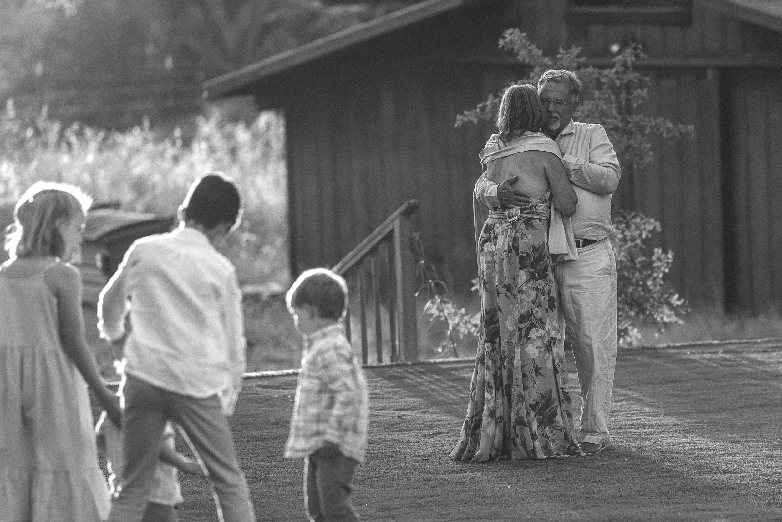Black and white photo showing people and onlookers gathered outside a wooden structure with sunlight filtering through trees.