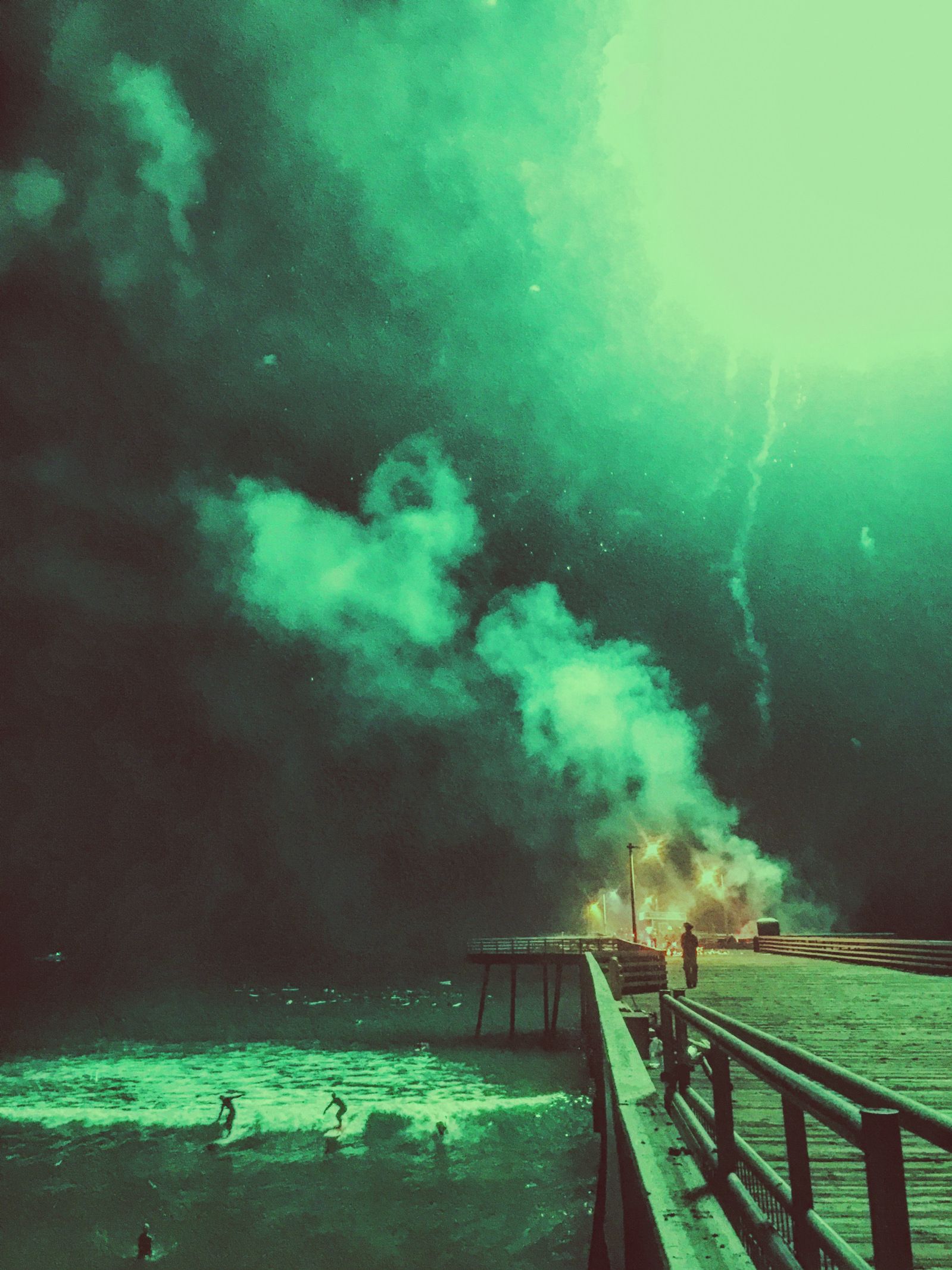 Surreal green smoke flares illuminate a pier extending into dark waters.