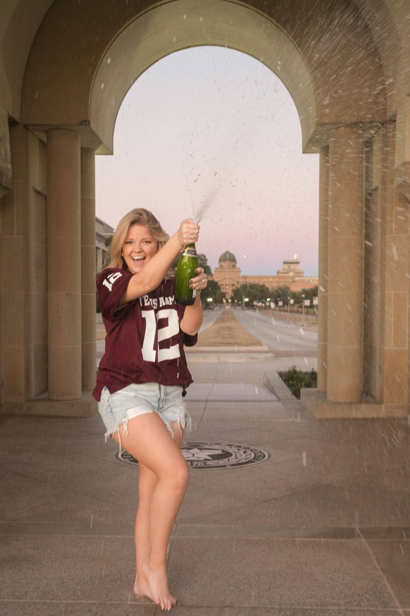Someone wearing a maroon #12 jersey and denim shorts poses under an arched colonnade at sunset.