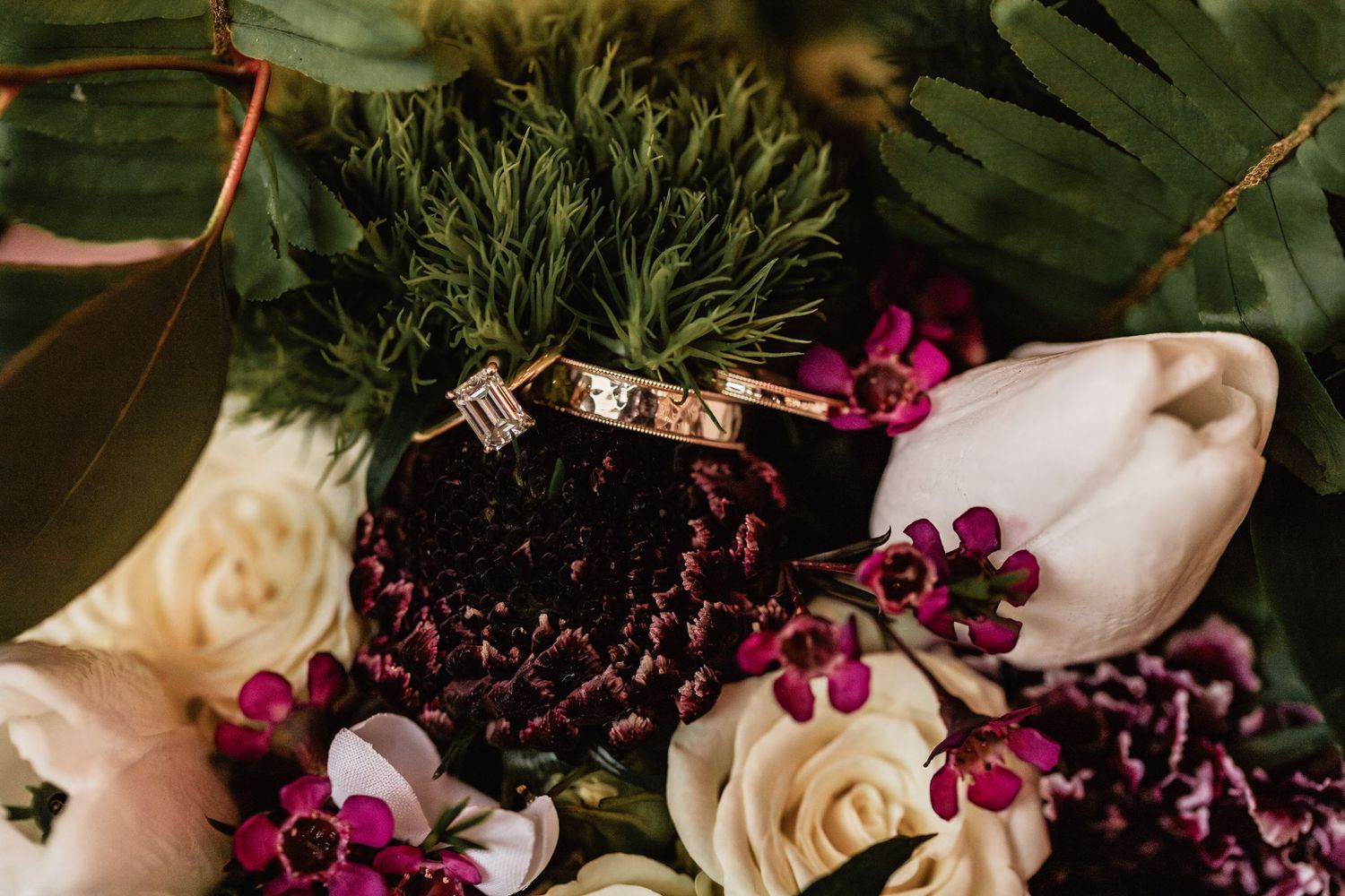 Close-up flat lay of gold wedding bands and emerald-cut diamond ring nestled in moody floral arrangement with roses, greenery, and tulips.