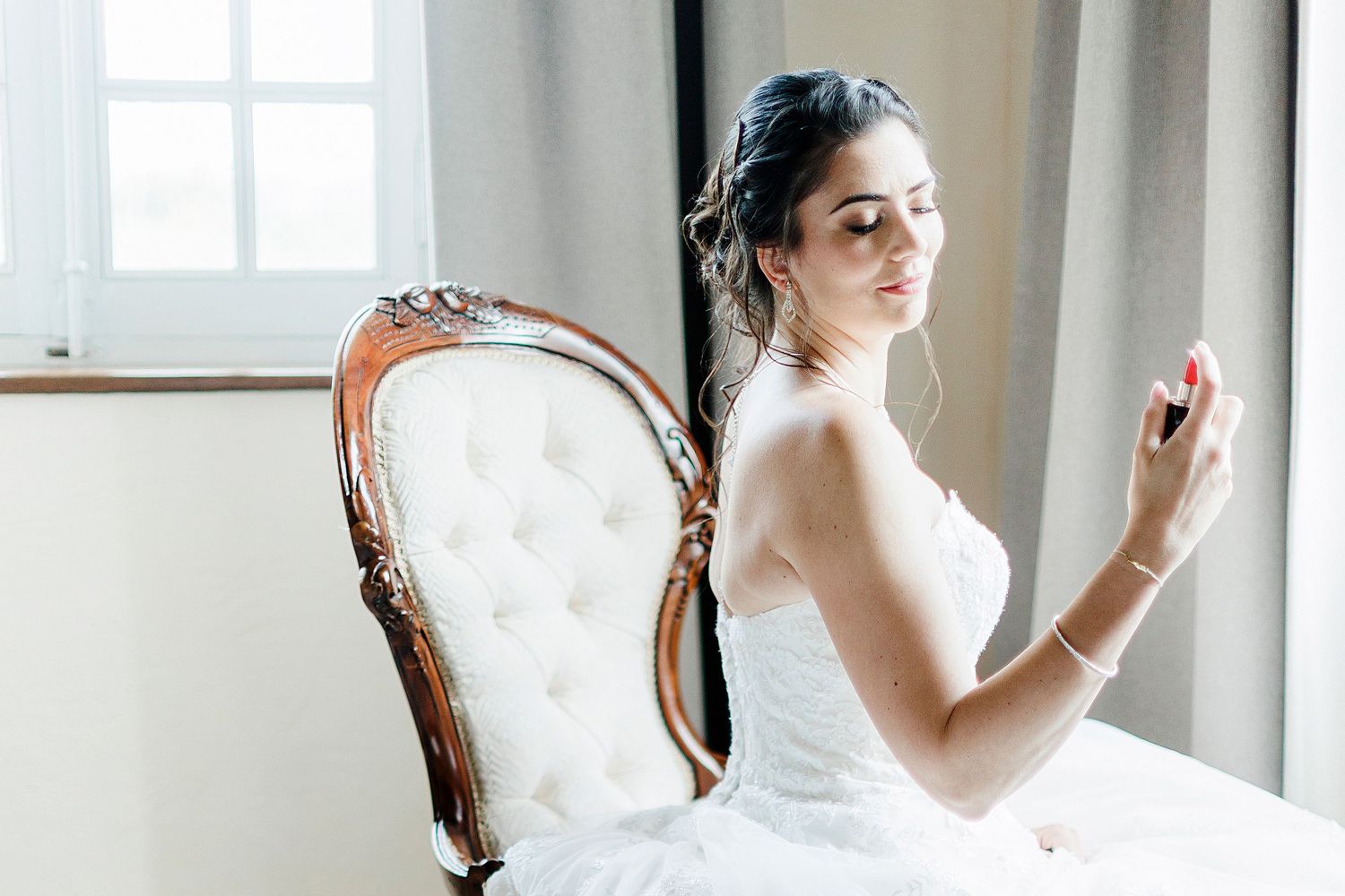 A bride in a white strapless wedding dress sits elegantly on an ornate vintage chair near a sunlit window.
