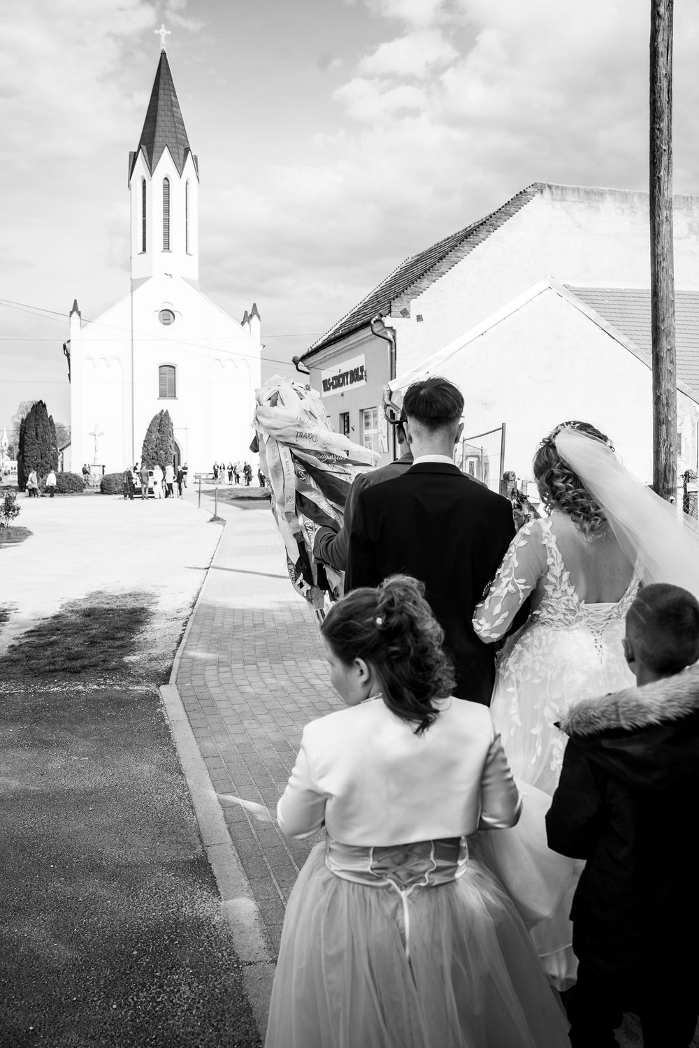 A black and white wedding procession walking down a pathway towards a church with a tall white steeple on a cloudy day.