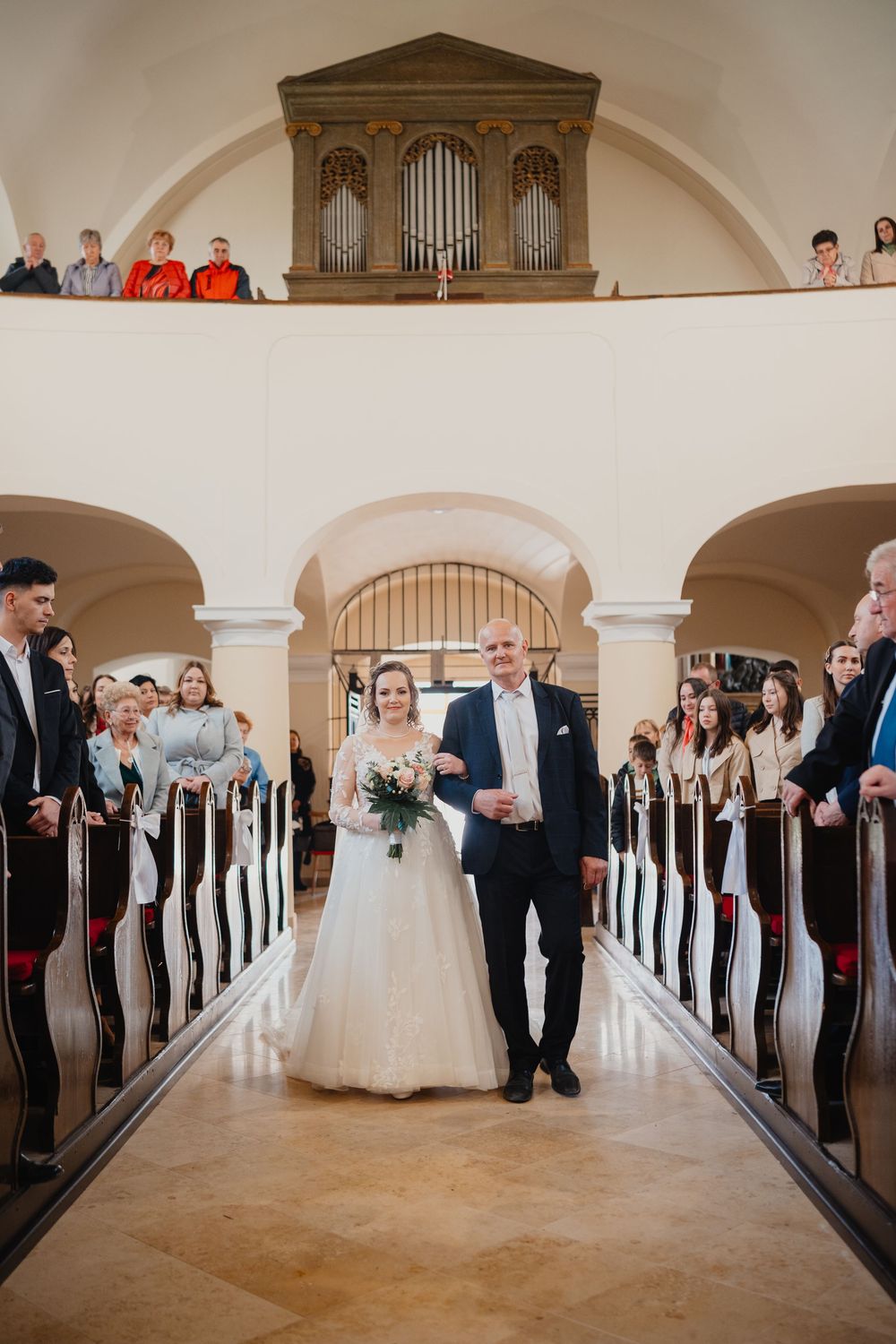 A bride walks down a church aisle lined with wooden pews beneath an ornate pipe organ during a traditional wedding ceremony.